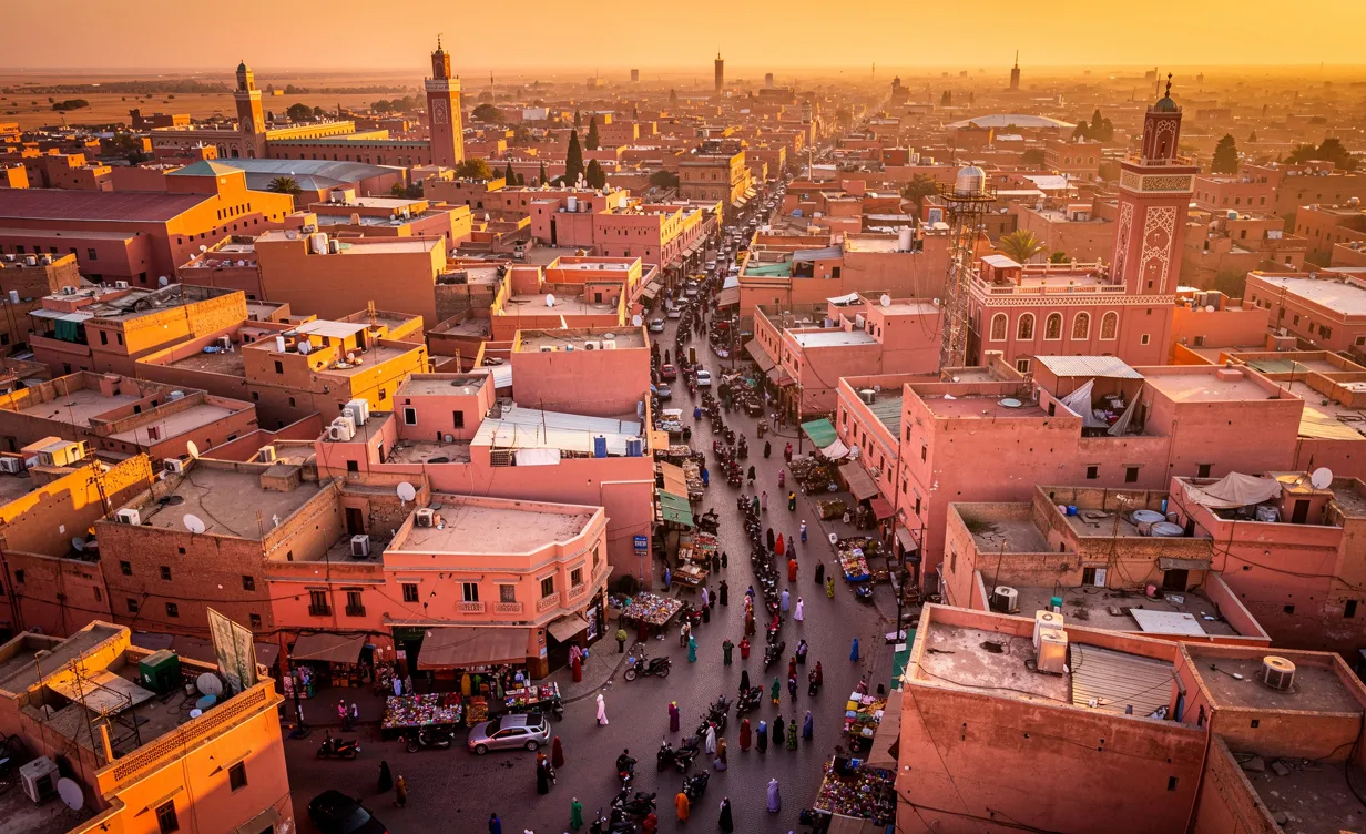 Vue aérienne photoréaliste de Marrakech avec ses toits en terre cuite, des minarets et des rues animées sous la lumière d’un coucher de soleil.