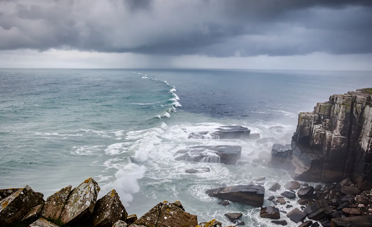 Une côte rocheuse où une ligne naturelle sépare les eaux bleu-vert de la Manche et celles plus sombres de la mer du Nord sous un ciel nuageux et une lumière brumeuse du matin.