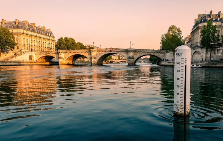 La Seine coule sous un pont en pierre à Paris, un repère de niveau d’eau partiellement immergé et les bâtiments historiques se reflètent dans l’eau au coucher du soleil.