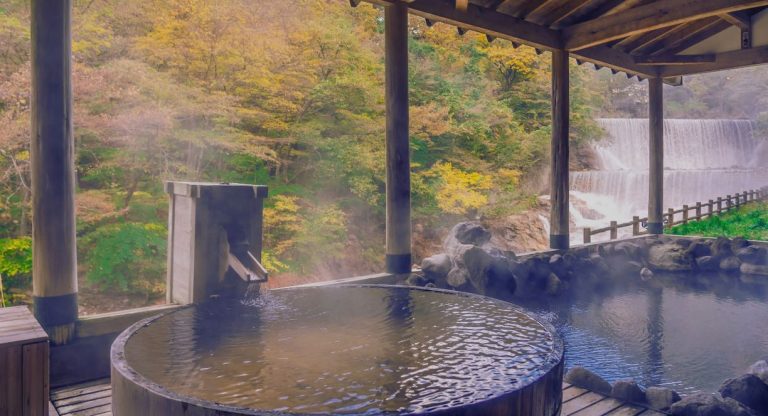 Onsen extérieur en bois avec eau chaude fumante entouré de feuilles dorées d'automne et d'une cascade en arrière-plan