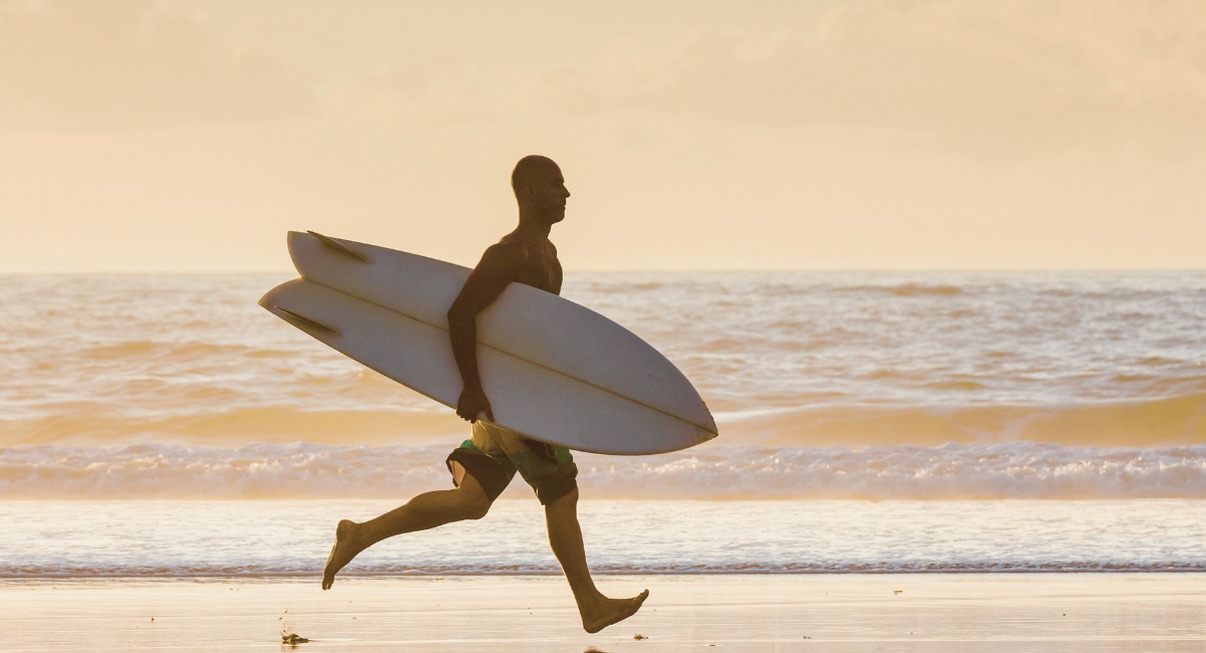 Surfeur en silhouette courant sur la plage avec sa planche de surf au coucher du soleil