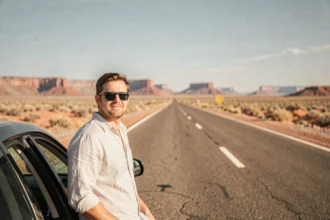 Homme appuyé contre une voiture sur une route désertique aux États-Unis, idéal pour illustrer un road trip en Amérique